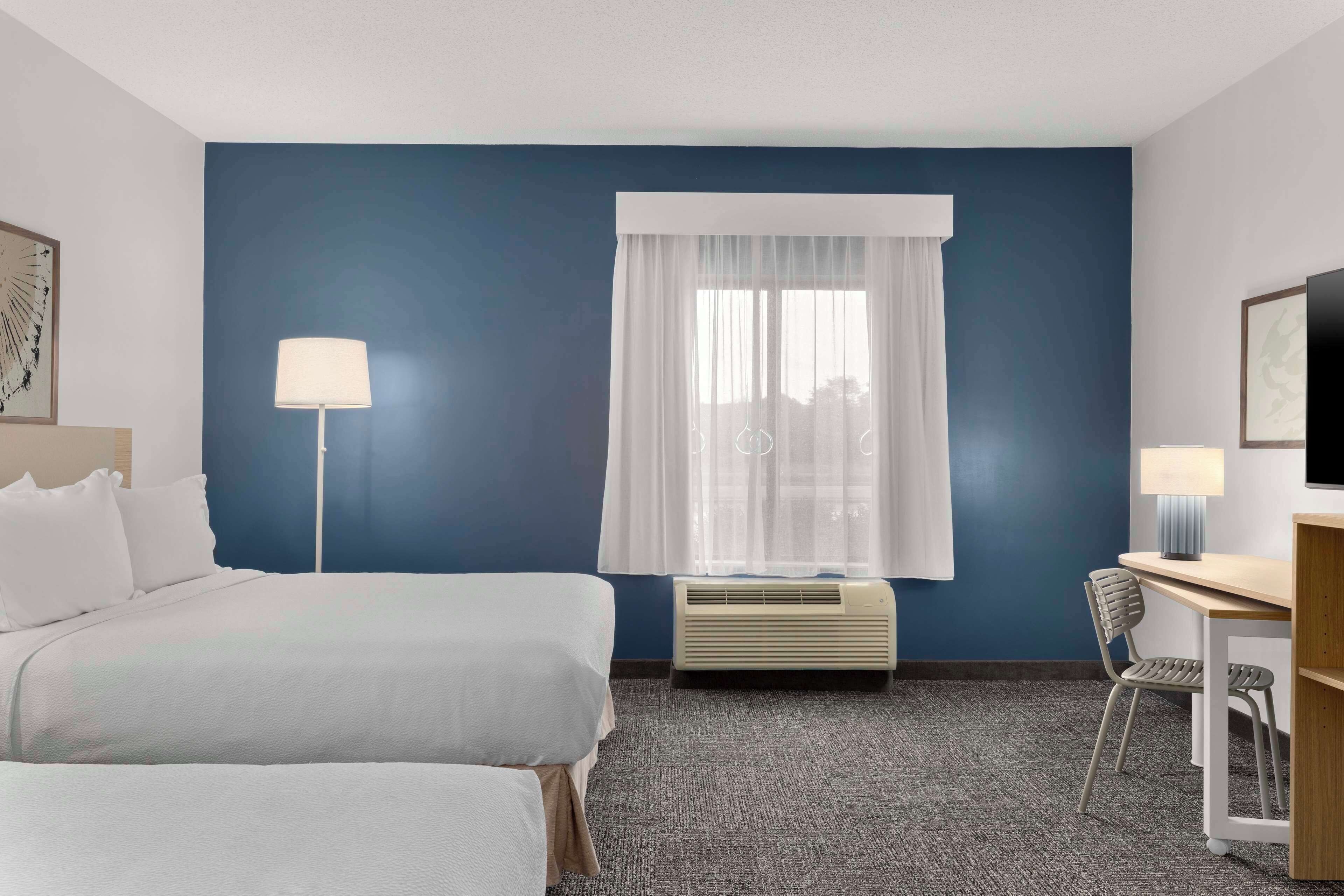 Une chambre d’hôtel comprenant un lit, un bureau, une chaise, une lampe, une fenêtre avec des rideaux légers et un appareil de climatisation.