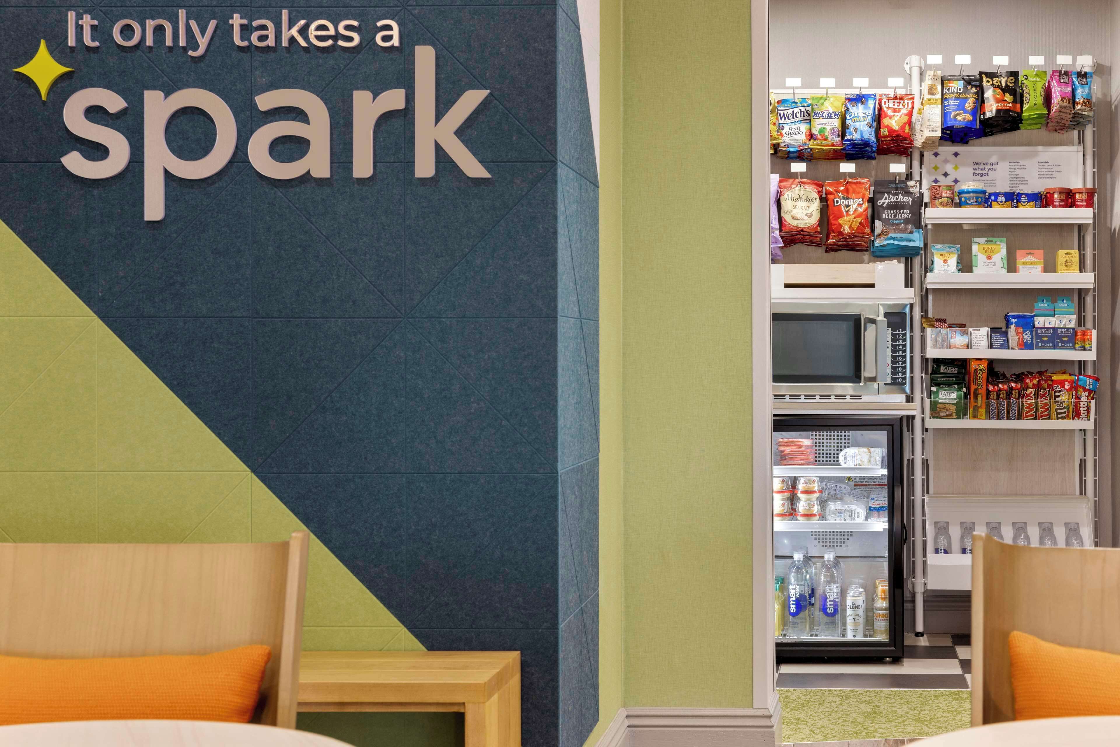 A hotel room with a bed, a small wooden table, a wall with a sign reading "It only takes a spark", and a mini-fridge stocked with beverages.