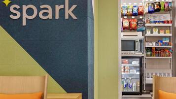 A hotel room with a bed, a small wooden table, a wall with a sign reading "It only takes a spark", and a mini-fridge stocked with beverages.
