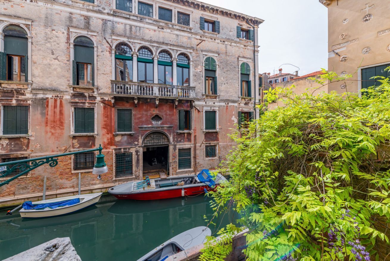 A canal with boats, a building with arched windows and balconies, and lush greenery.