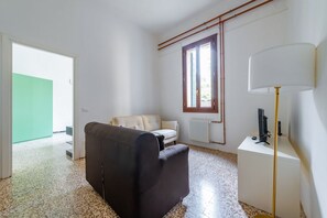 A room with a sofa, a lamp, a radiator, a window, and a door leading to a green area.