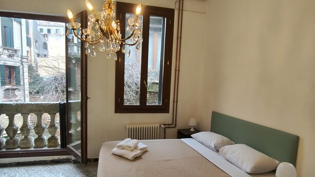 A bedroom with a bed, a nightstand, a lamp, a chandelier, a window with a view of buildings, and a radiator. - GuestHost - Apartment located on the first floor of an ancient building without a lift.Splendid view of the canal from where the typical Venetian gondolas pass.It accommodates 4 people in 85 square meters of space with two double bedrooms.We are located  (Venezia)