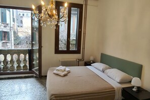 A bedroom with a bed, a nightstand, a lamp, a chandelier, a window with a view of buildings, and a radiator.