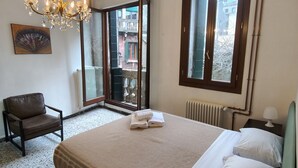 A bedroom with a bed, a chair, a chandelier, a radiator, and a window with a view of buildings. - GuestHost - Apartment located on the first floor of an ancient building without a lift.Splendid view of the canal from where the typical Venetian gondolas pass.It accommodates 4 people in 85 square meters of space with two double bedrooms.We are located  (Venezia)