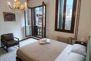 A bedroom with a bed, a chair, a chandelier, a radiator, and a window with a view of buildings.