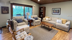 A spacious living room with a wooden ceiling, a large window with a view of greenery, a beige sofa set, a wooden coffee table, and a bookshelf. - Seneca Lake View Retreat sits secluded on a 2 acre lot. (Himrod)
