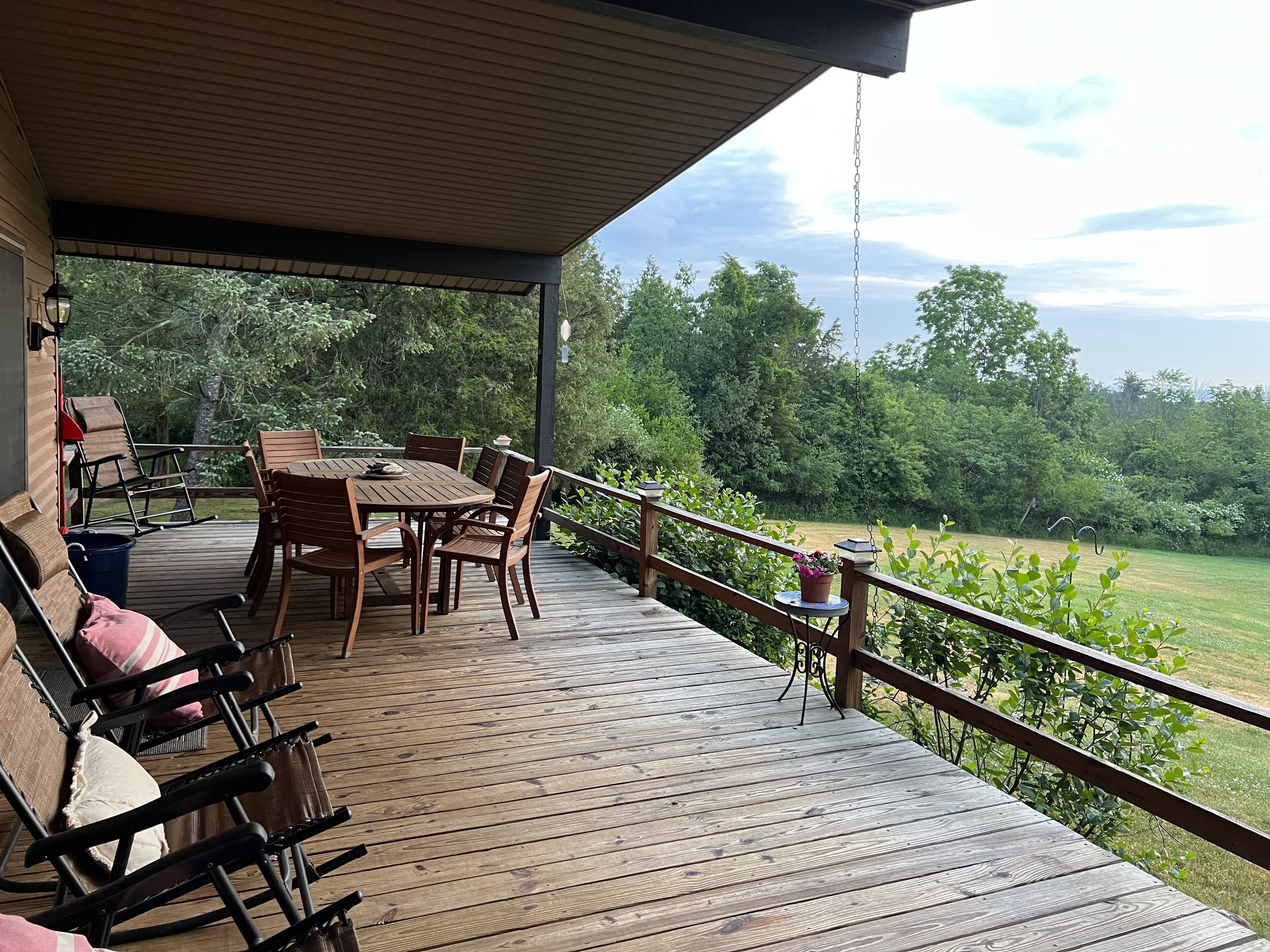 A wooden deck with a table and chairs, a hanging light fixture, and a view of a green landscape.
