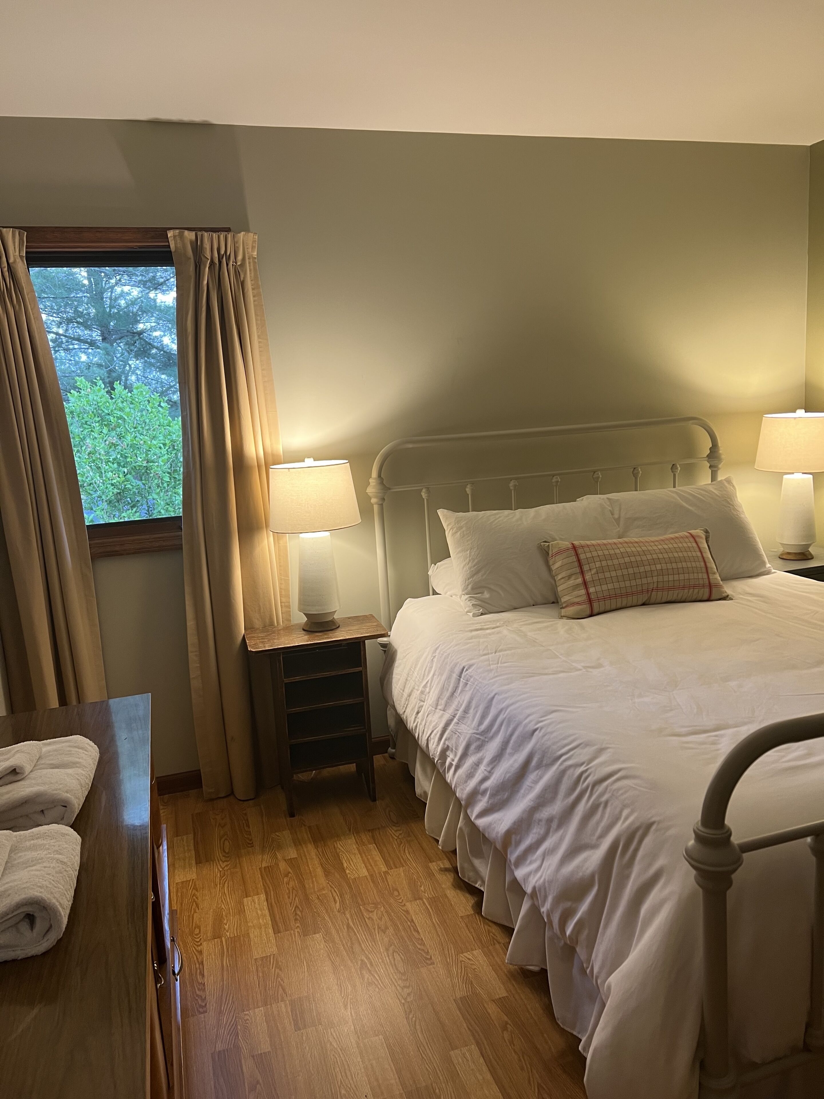 Een slaapkamer met een bed, twee nachtkastjes met lampen, een houten nachtkastje, een raam met gordijnen en een houten vloer.