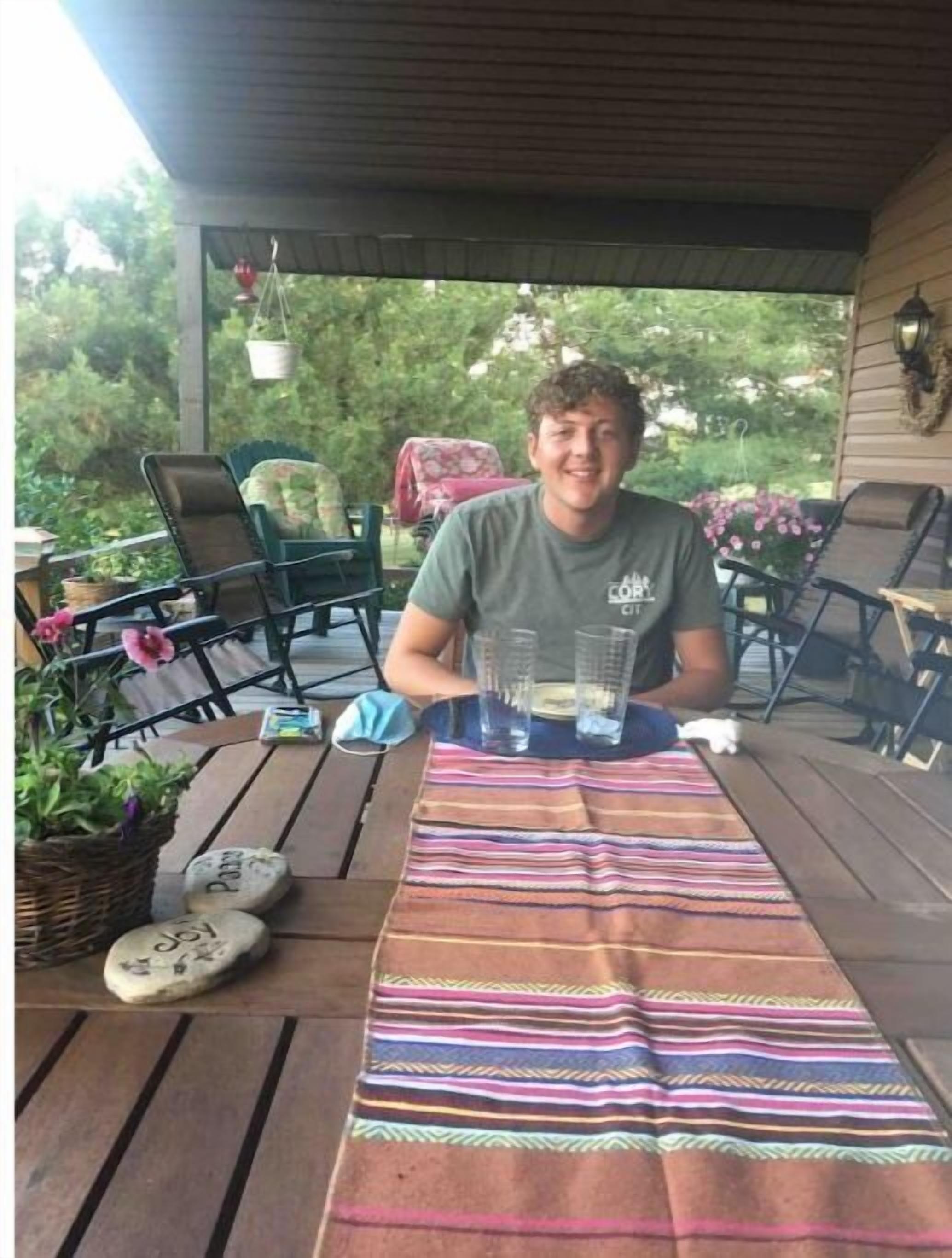 Une personne est assise à une table en bois recouverte d’une nappe rayée aux couleurs vives, entourée de chaises et de plantes en pot.