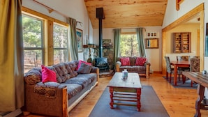 A cozy cabin interior with a wooden ceiling, large windows, a stone fireplace, a sofa, a table, and a dining area. - Rustic Pagosa Springs Cabin w/ Hot Tub + Game Room (Pagosa Springs)