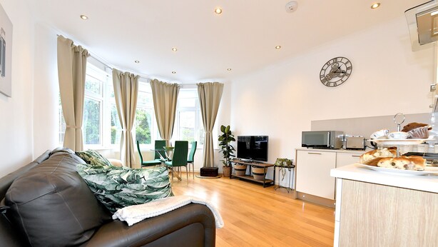 Een moderne woonkamer met een leren bank, een eethoek met tafel en stoelen, een keuken met een televisie en een klok aan de muur.