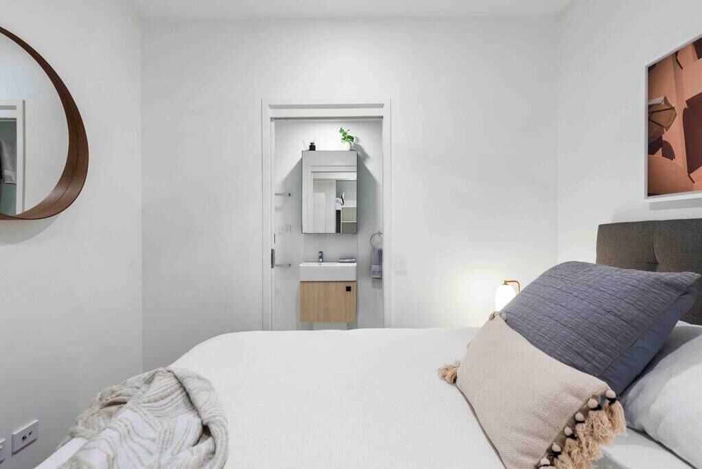 A modern bedroom with a bed, a bedside table, a nightstand, a mirror, a door leading to a bathroom, and a wall-mounted artwork.