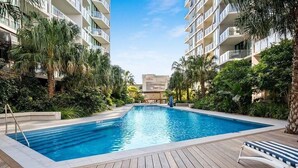 A swimming pool with a wooden deck, surrounded by palm trees and greenery, with high-rise buildings in the background.