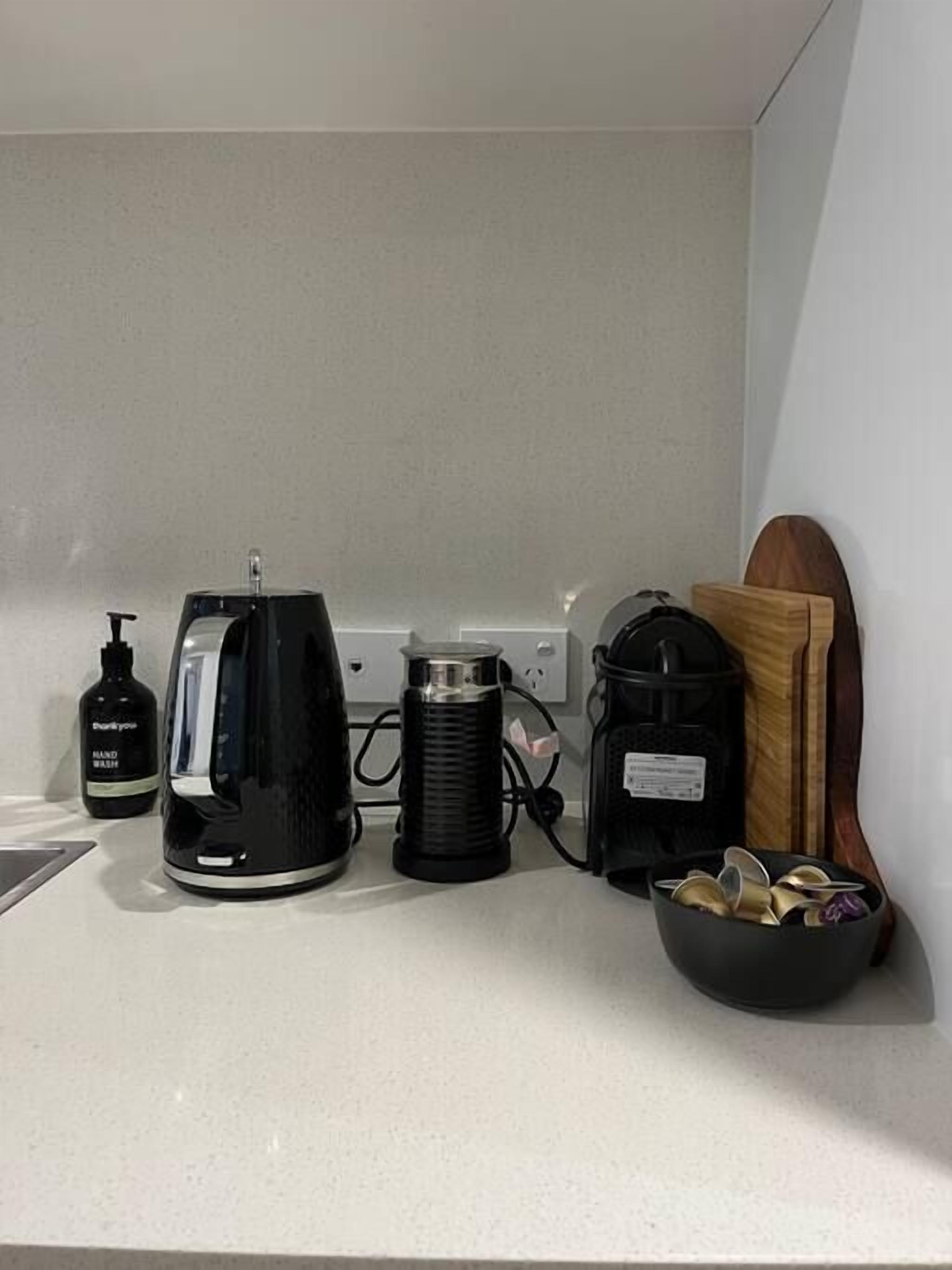Una encimera de cocina con cafetera, hervidor, dispensador de jabón, regleta de enchufes y un cesto de latas.