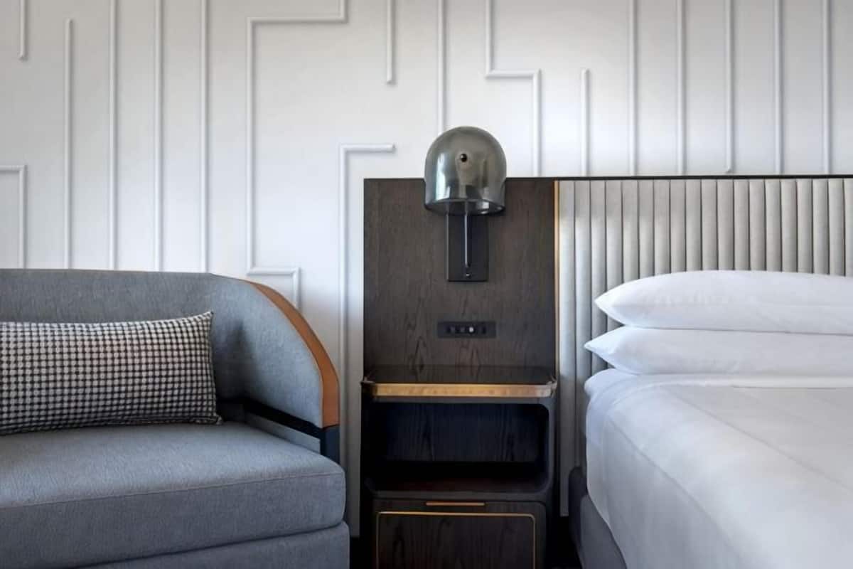 Una habitación de hotel moderna con un sofá gris, una mesita de noche de madera con una lámpara y una cama bien hecha.