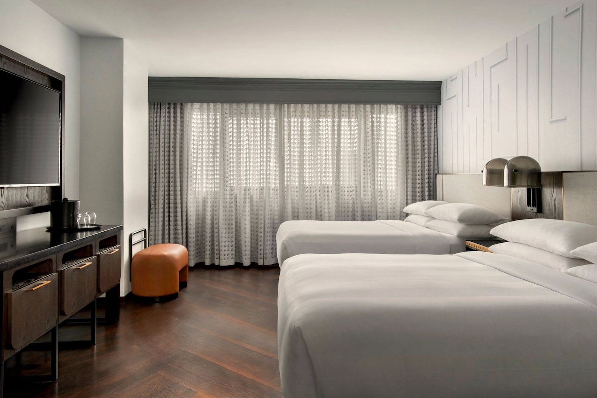 A hotel room with a bed, a desk with a flat-screen TV, a small stool, and a window with sheer curtains.