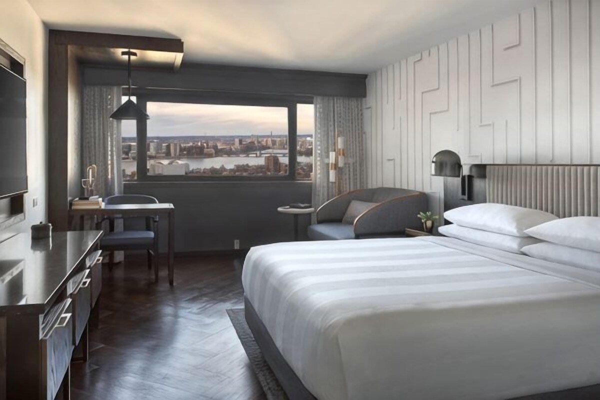 A hotel room with a bed, a desk, a chair, a window with a city view, and a cityscape visible through the window.