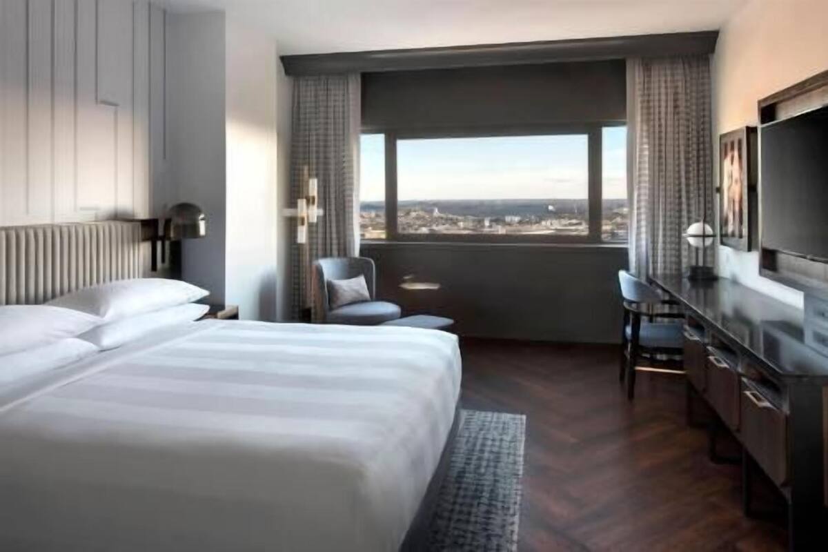 Habitación de hotel con cama, escritorio, silla, televisor y una ventana grande con vistas a la ciudad.