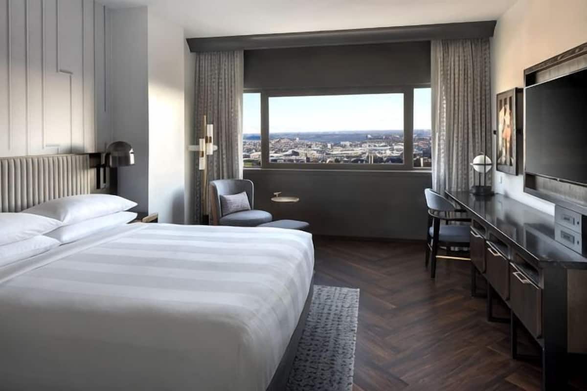 Habitación de hotel con una cama grande, un escritorio, una silla, un televisor y vistas a un paisaje urbano.