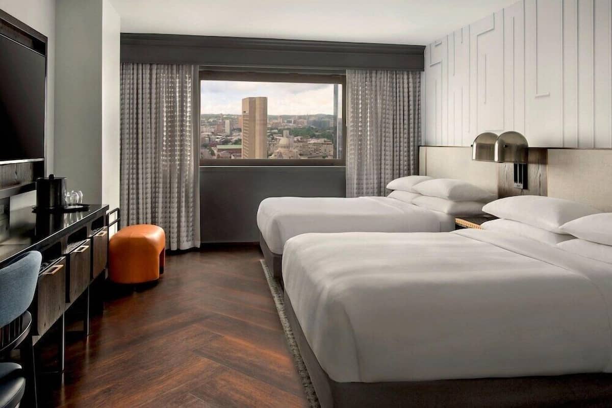 Habitación de hotel con dos camas, televisión, escritorio y vistas a un paisaje urbano.