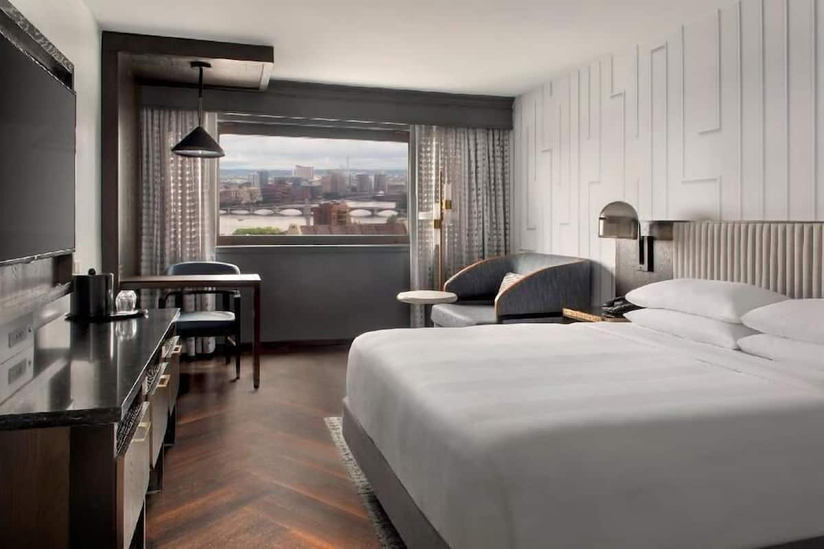 Habitación de hotel con una cama grande, un escritorio, una silla, una mesita y vistas a un paisaje urbano a través de una ventana.