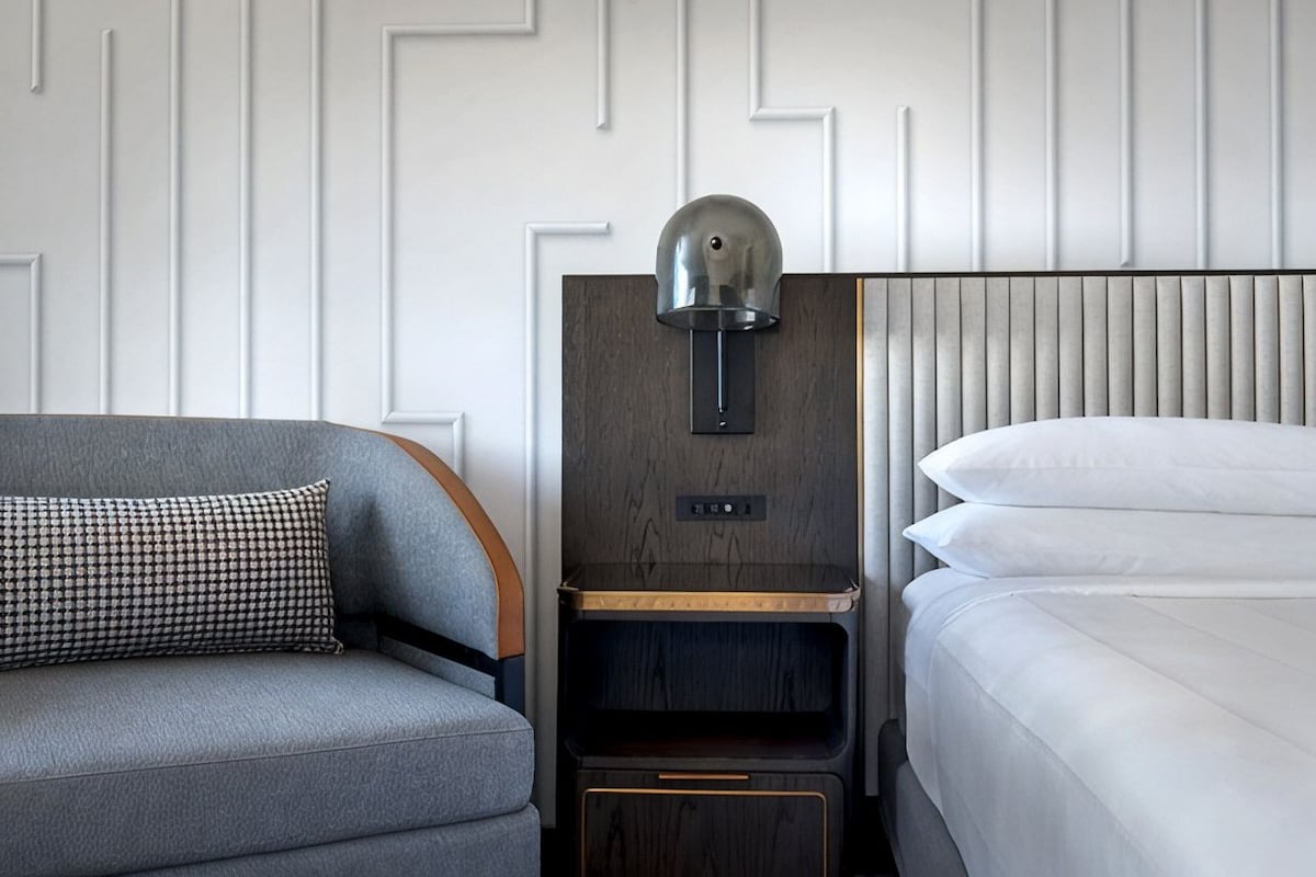 A hotel room with a grey sofa, a bedside table with a lamp, and a bed with white bedding.