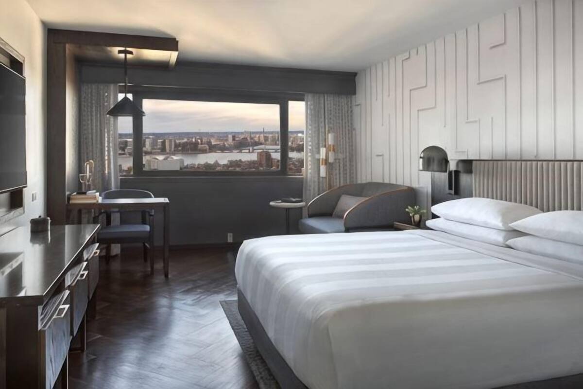 Habitación de hotel con cama, escritorio, silla y ventana que ofrece vistas a la ciudad, con un paisaje urbano visible a través de ella.