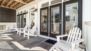 A covered outdoor seating area with Adirondack chairs and tables, a wooden deck, and a black-framed glass door leading to the interior.