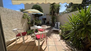 Een terras buiten met witte stoelen en een tafel, een geribbelde parasol en diverse planten.