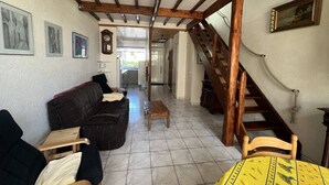 Een ruime woonkamer met een donkerbruine bank, een houten salontafel en een gele tafelkleed met dessin. Een houten trap met touwbalustrade leidt naar een verdieping.
