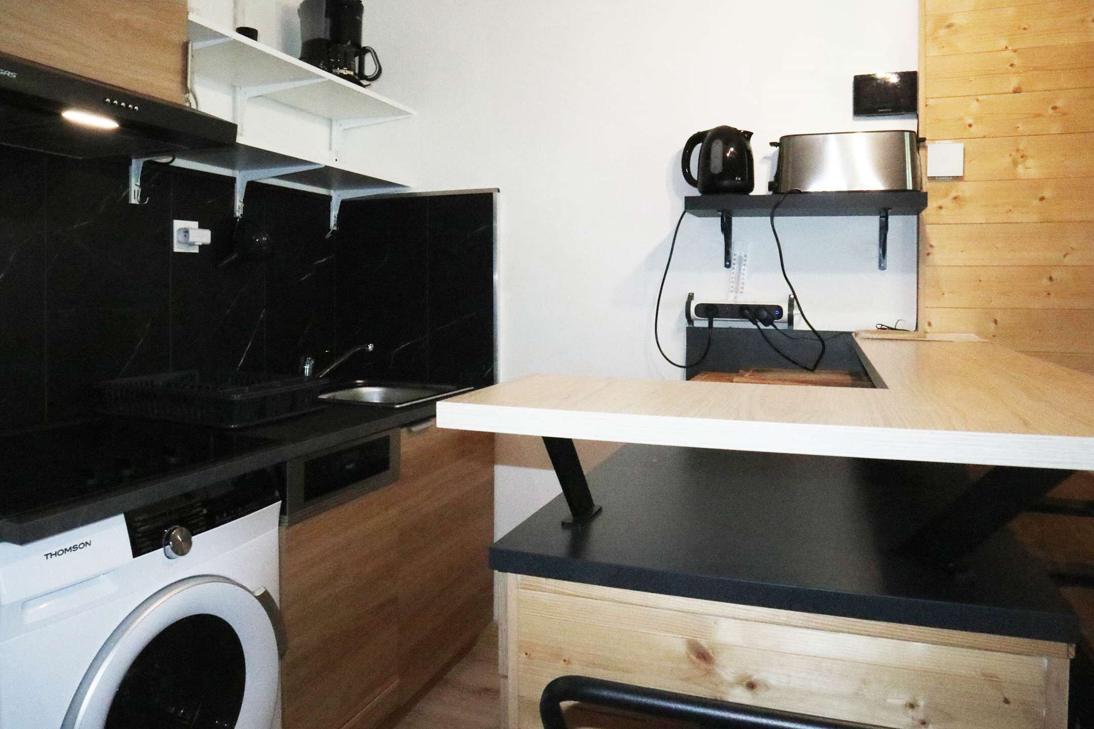 Une cuisine compacte dotée d’un lave-linge intégré, d’un plan de travail en bois et d’une étagère sur laquelle se trouvent une bouilloire et une casserole.