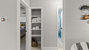Corredor que dá para um armário com prateleiras, uma cama e uma mala no chão.