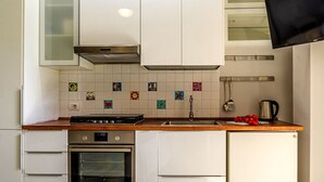 A kitchen with white cabinets, a stainless steel oven, a sink, and a small refrigerator. There are colorful tiles on the backsplash and a flat-screen TV mounted above the sink. - Perla Marina-Giardini Naxos (Giardini Naxos)