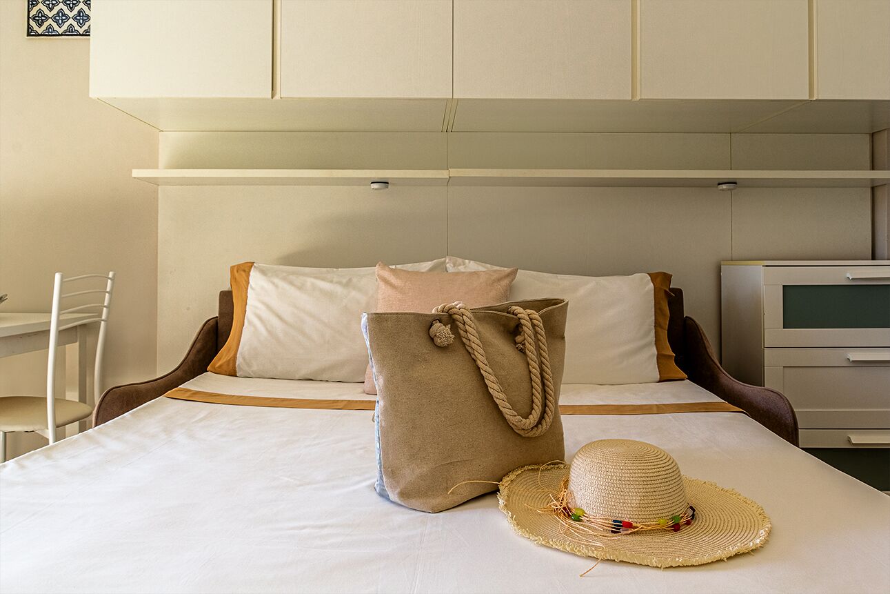 Siististi pedattu sänky, jonka päällä on beige väriin sävyttynyt laukku ja olkihattu.
