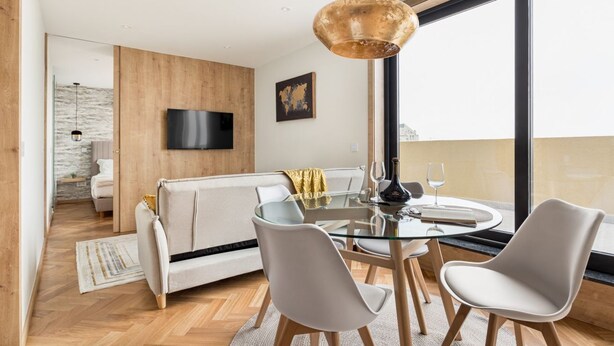 A modern living room with a glass dining table, white chairs, a sofa, and a large window with a view.