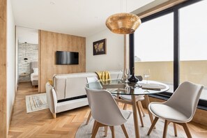 Una sala de estar moderna con una mesa de comedor de cristal, sillas blancas, un sofá y un amplio ventanal con vistas.