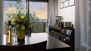 Una mesa de comedor con una botella de vino, dos copas y un florero con flores. Se ve un paisaje de árboles y una puesta de sol a través de una ventana.