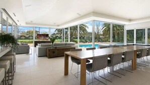 A spacious indoor area with a large dining table, several chairs, and a sofa. There is a pool visible through large windows. - Tanunda House - Luxury 5 BR in Barossa Wine Region (Tanunda)