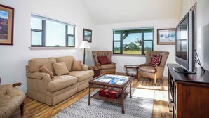A living room with a sofa, two armchairs, a coffee table, a TV, and large windows with a view of trees.