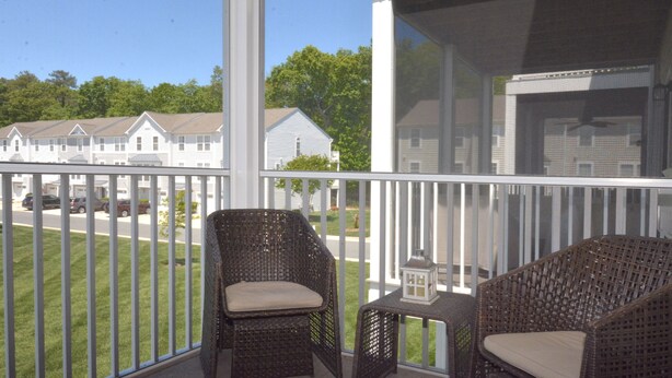 A balcony with wicker chairs and a small table, overlooking a street with houses and a clear sky. - Rehoboth Beach Townhouse Pool Parking Porch (Rehoboth Beach)