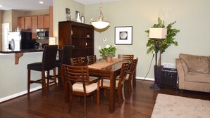 A dining area with a wooden table and chairs, a kitchen in the background, and a living room with a couch and a lamp. - Rehoboth Beach Townhouse Pool Parking Porch (Rehoboth Beach)