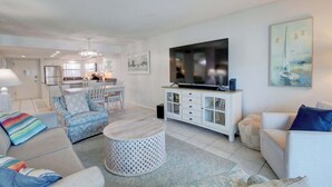 Een moderne woonkamer met een televisie, bank, fauteuils, een eethoek en een keuken.