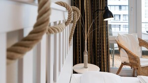 Una moderna camera d'albergo con un letto bianco, un tavolo rotondo, una poltrona in vimini, una grande finestra con vista sugli edifici e un decoro in corda al muro.