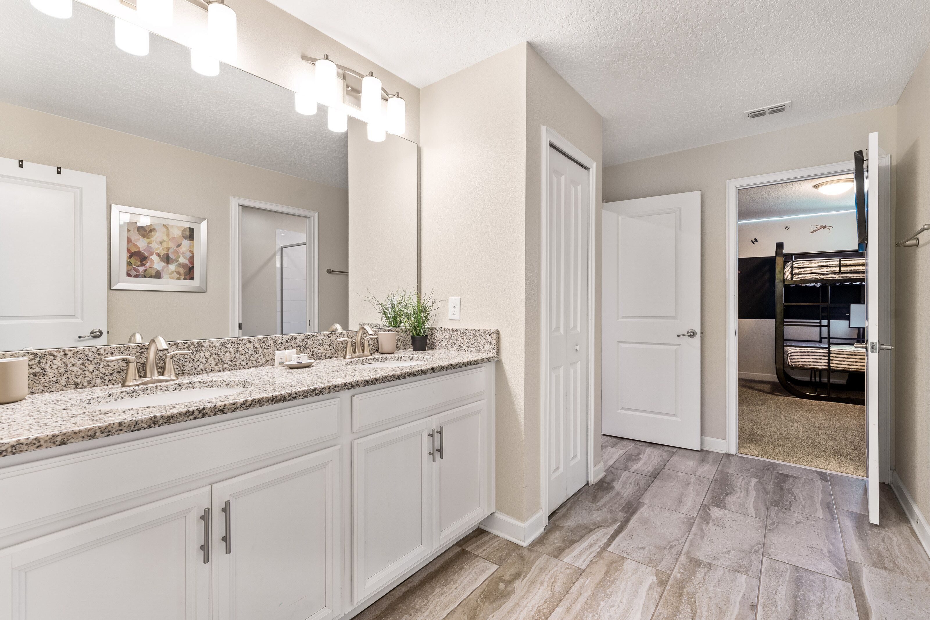 Un baño moderno con encimera de granito, muebles blancos, un espejo grande y una zona de ducha visible a través de una puerta abierta.