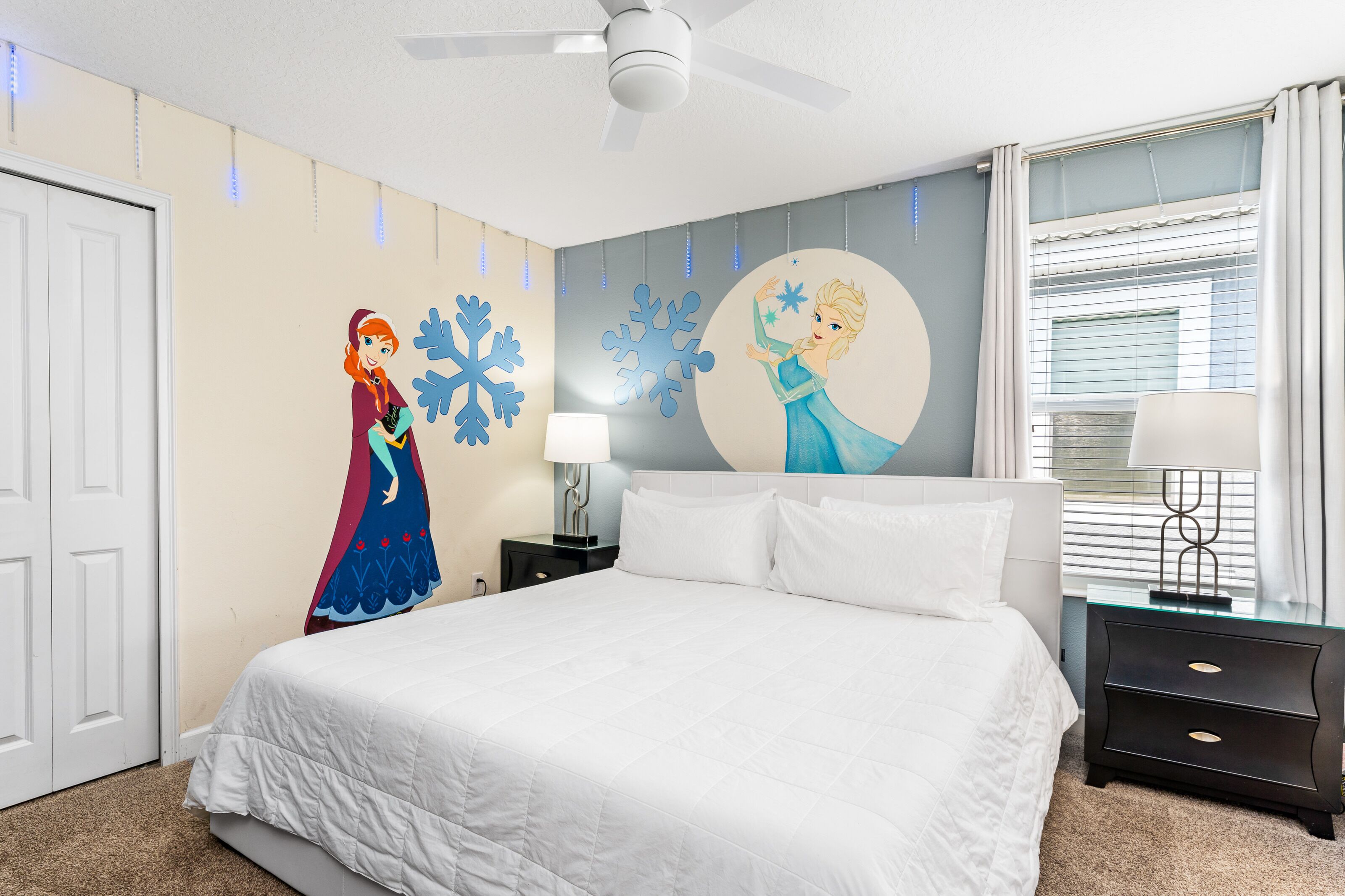 A bedroom with a bed, a nightstand, a lamp, a ceiling fan, and wall decorations including a character and snowflakes.