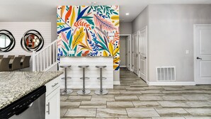 Eine Küche mit Granit Arbeitsplatte, weißen Schränken und einem farbenfrohen, abstrakten Wandbild. Drei Barhocker und eine weiße Tür sind im Hintergrund zu sehen.