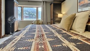 Quarto de hotel com uma cama grande, um ventilador, uma cadeira, uma janela com vista para montanhas e um quadro na parede.