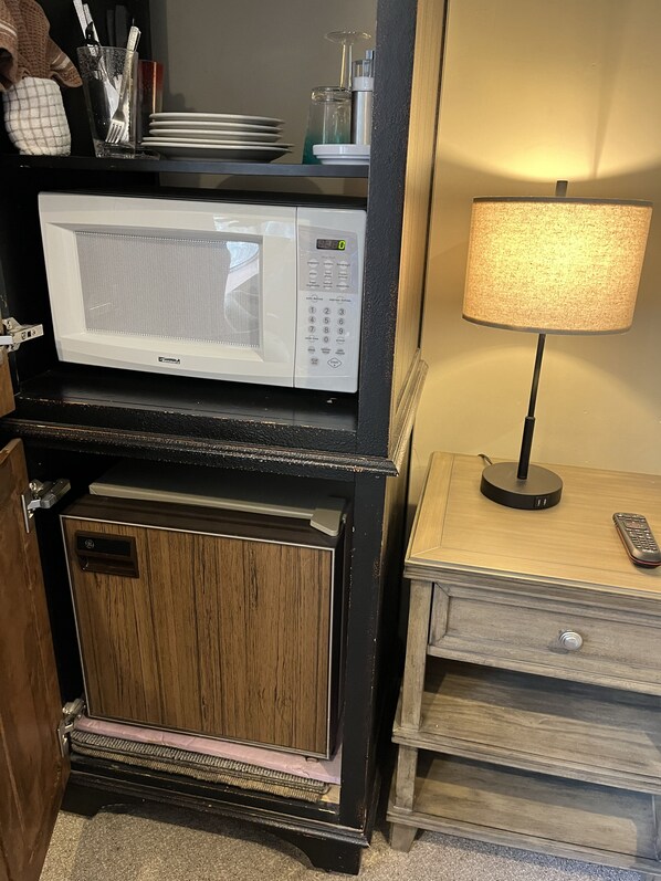 A compact kitchen with a microwave, a small refrigerator, and a lamp on a bedside table.