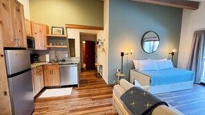 Habitación de hotel pequeña con kitchenette, una cama con colcha azul estampada, un espejo redondo y suelo de madera.