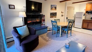 A living room with a fireplace, a television, a dining table with chairs, and a kitchen area with wooden cabinets. - Suite Situation in the Ski Village (Taos Ski Valley)